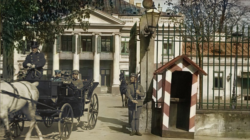 A carriage with two passengers leaving through a guarded gate with a booth; in the background, a building with a colonnade