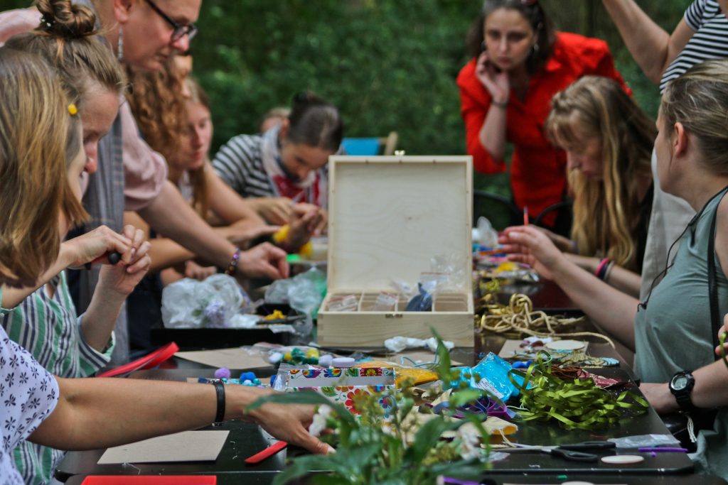 In the centre, a table with various items used in handicrafts, e.g. boxes, ribbons and strings. Workshop participants sit or stand around the table.