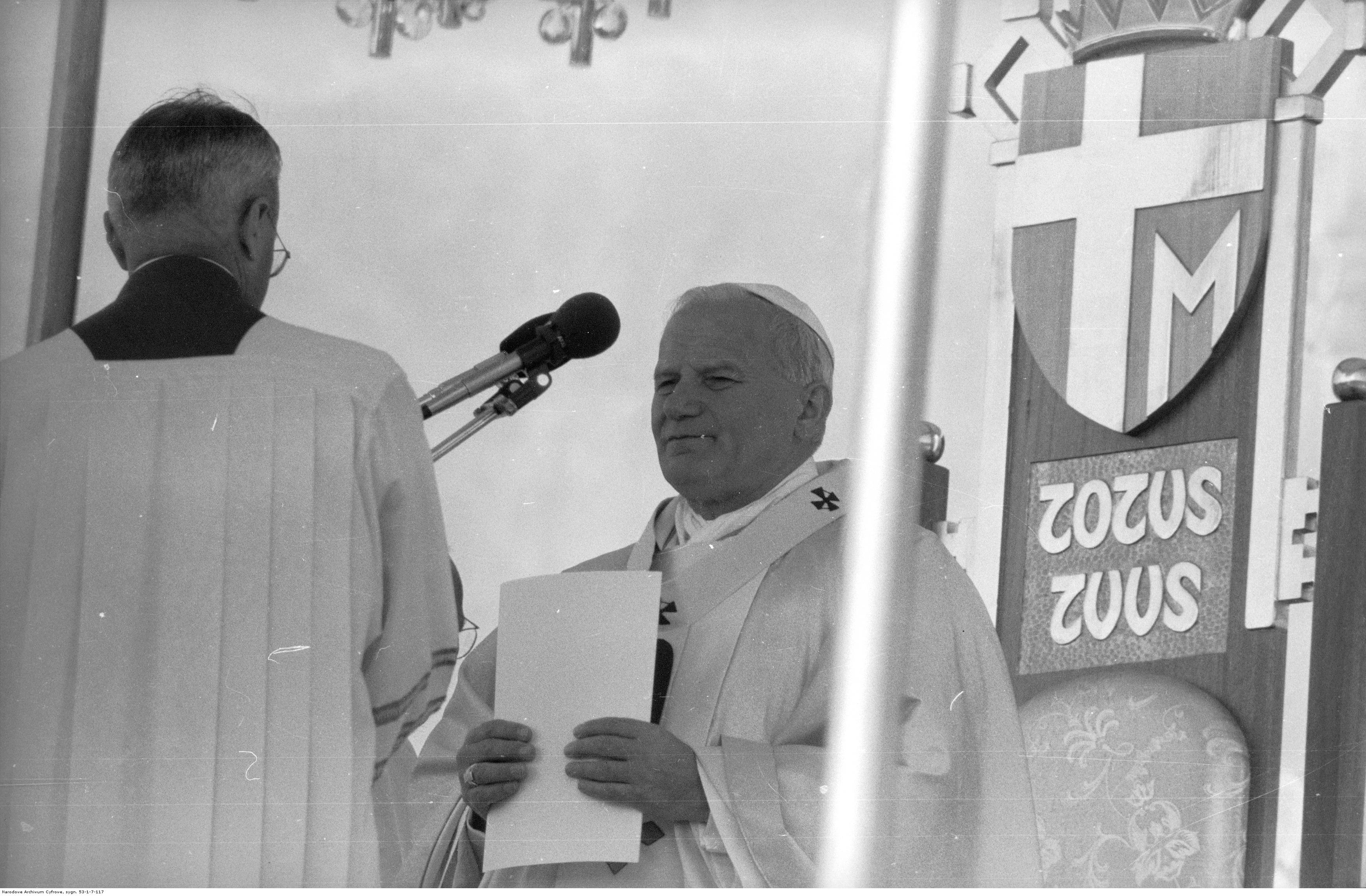 W centrum Jan Paweł II przygotowuje się do homilii. Z lewej widoczny ceremoniarz papieski, ks. prał. Virgilio Noe przygotowujący mikrofon. Z prawej widoczny herb papieża Jana Pawła II jako element tronu papieskiego.