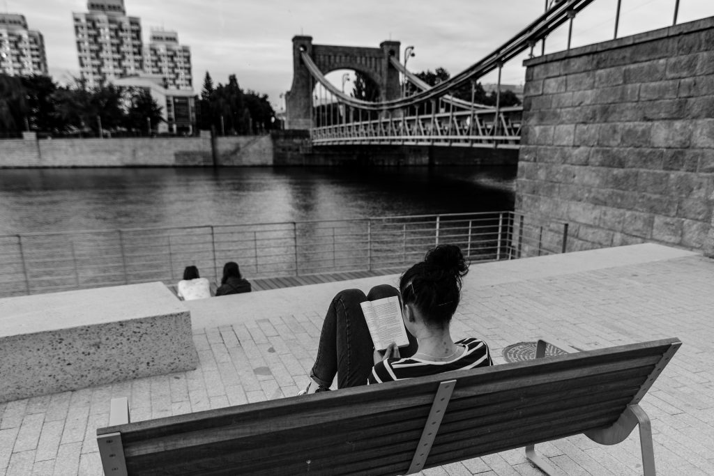 na pierwszym planie kobieta tyłem, czytająca na ławce książkę, w tle, za rzeką, wieżowce mieszkalne z okresu PRL