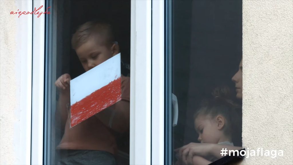chłopczyk w mieszkaniu przykleja ręcznie wykonaną flagę polski do okna, obok asystują mu mama i siostra
