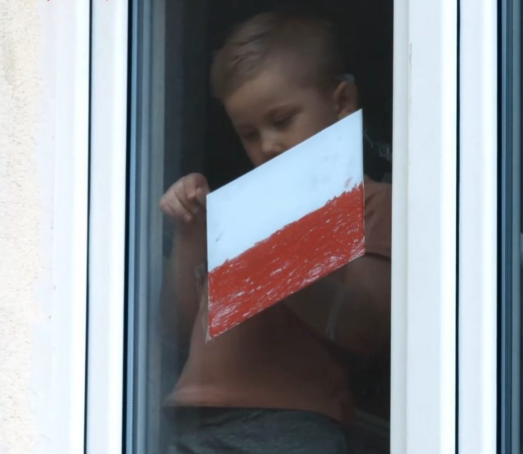 chłopczyk w mieszkaniu przykleja ręcznie wykonaną flagę polski do okna