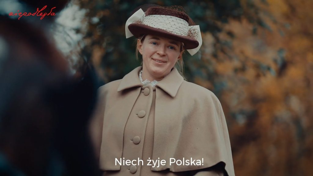 kobieta na powozie w retro ubraniu patrzy w dół z uśmiechem po wypowiedzeniu kwestii niech żyje polska