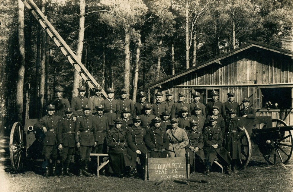 grupa żołnierzy w mundurach w fotografii pozowanej z dwoma działami drabiną i koszarami i lasem w tle