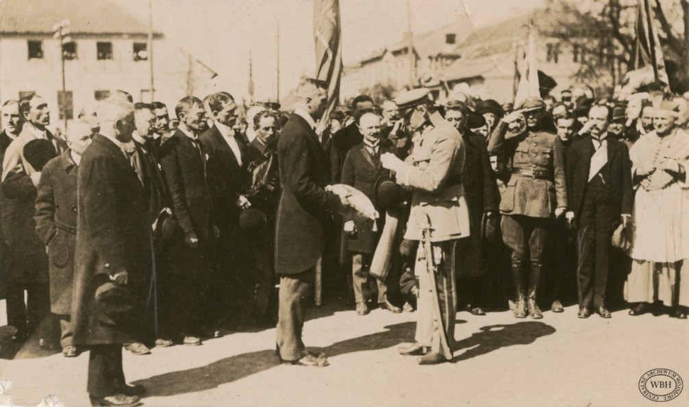 przed tłumem zgromadzonym na rynku uroczysty sztandar, a przed nim dwóch mężczyzn - po lewej trzyma poduszkę z symbolami miasta, po prawej Piłsudski do poduszki przypina order