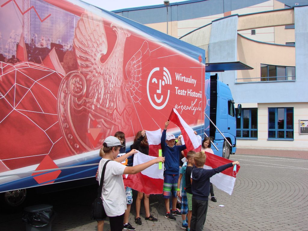 grupa dzieci z biało-czerwonymi flagami przed autobusem WTH, w tle budynek MOSiR
