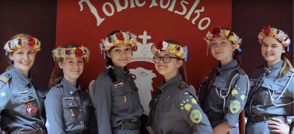 sześć dziewczyn w mundurach harcerskich i wiankach opolskich na głowie na tle baneru Tobie Polsko