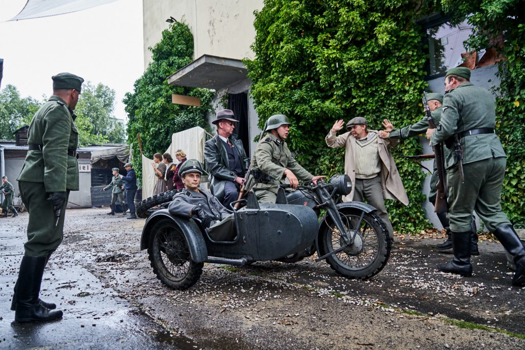 łapanka, na pierwszym planie naziści w mundurach na motocyklu z doczepką, w tle cywile ustawieni w rzędzie z rękami na głowie, dookoła rozproszeni żołnierze