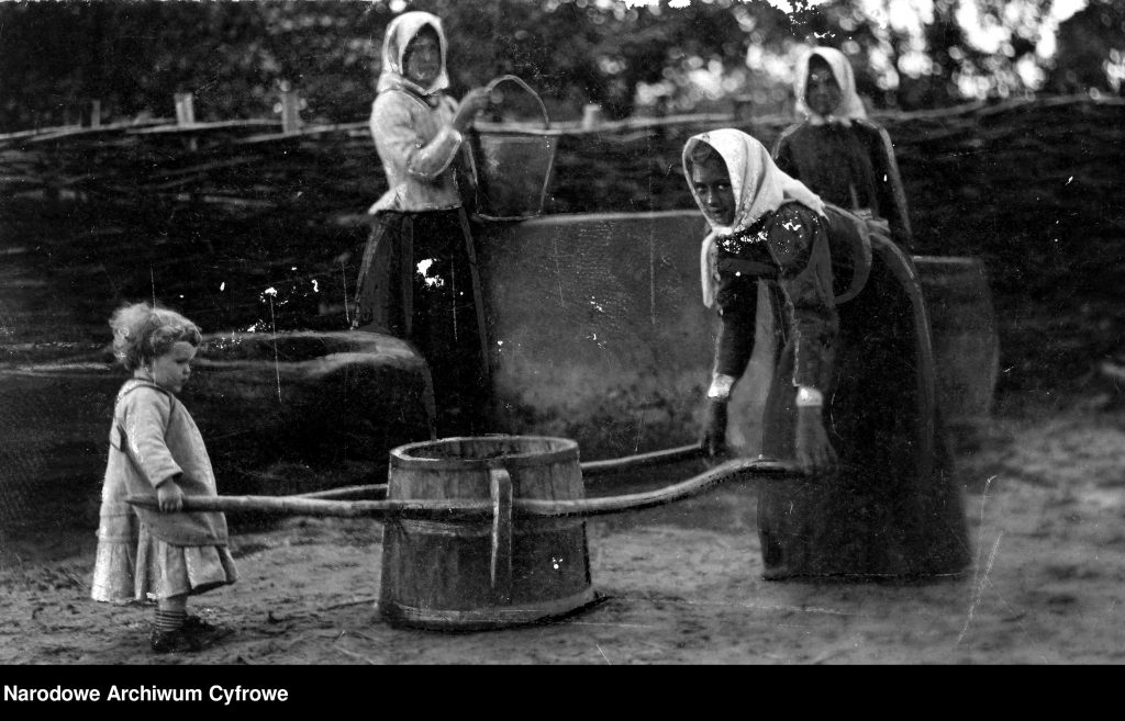 Kobiety z dzieckiem przy wiejskiej studni, dziecko i kobieta łapią wiadro na drągach