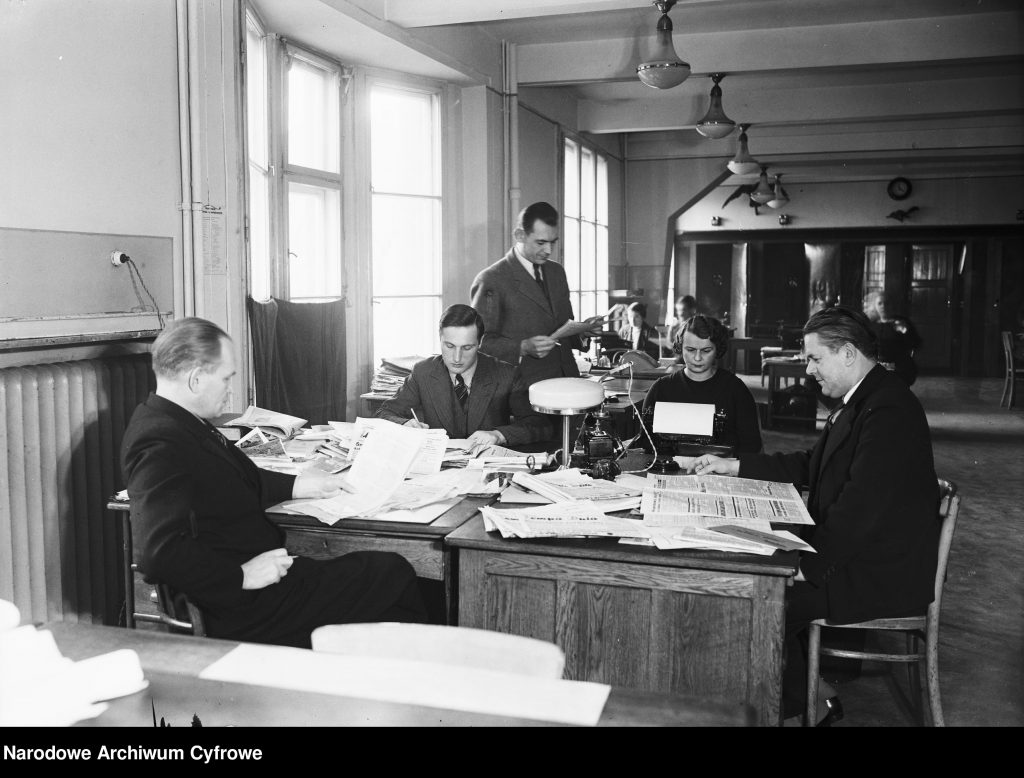 biuro, w środku biurka zarzucone papierami, wśród nich rozsadzeni mężczyźni przy pracy