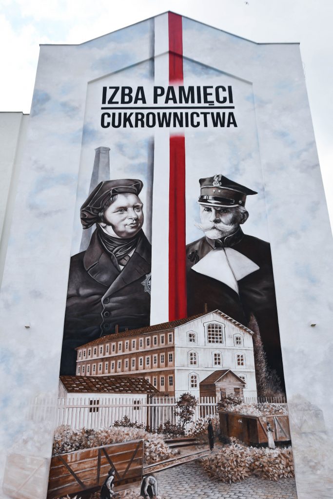mural złożony z dwóch historycznych postaci iddzielonych od siebie biało-czerwoną wstęgą, pod nimi widoczny namalowany budynek cukrowni
