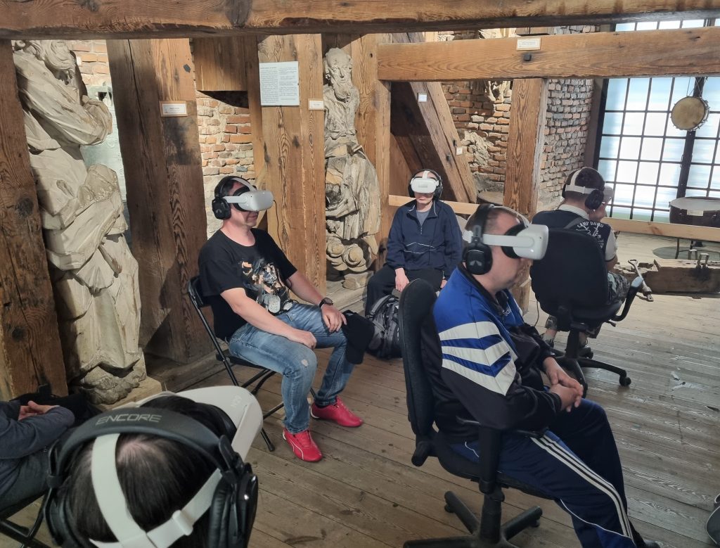 ludzie siedzący na obrotowych fotelach zzałożonymi googlami VR i słuchawkami, sala ma historyczny wystrój z belek drewnianych i cegieł