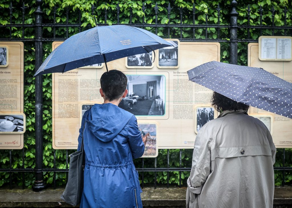 wystawa zawieszona na płocie, przed nią widzowie pod parasolami