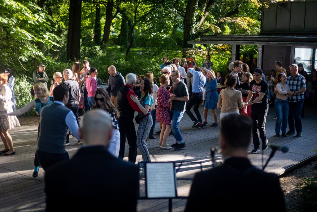 na pierwszym planie muzycy siedzącu z nutami, przed nimi ludzie tańczący na parkiecie