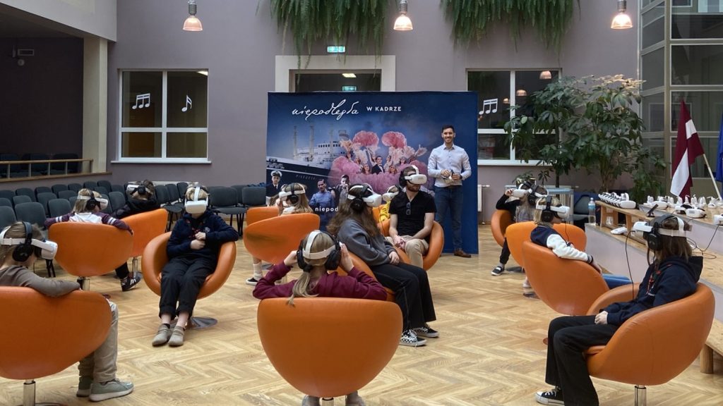dzieci na obrotowych fotelach oglądają film VR, w tle ścianka z nazwą projektu