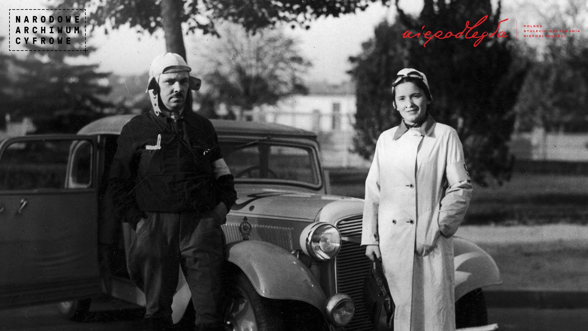czarno-białe zdjęcie przedstawiające mężczycnę i kobietę w latach 30-tych XXw. w czapkach i goglach dla kierowców na tle samochodu z epoki.