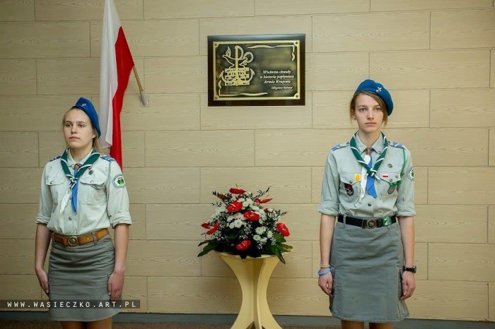 harcerki stoją pod tablicą pamiątkową, na ścianie wiszą biało-czerwone flagi i kwiaty