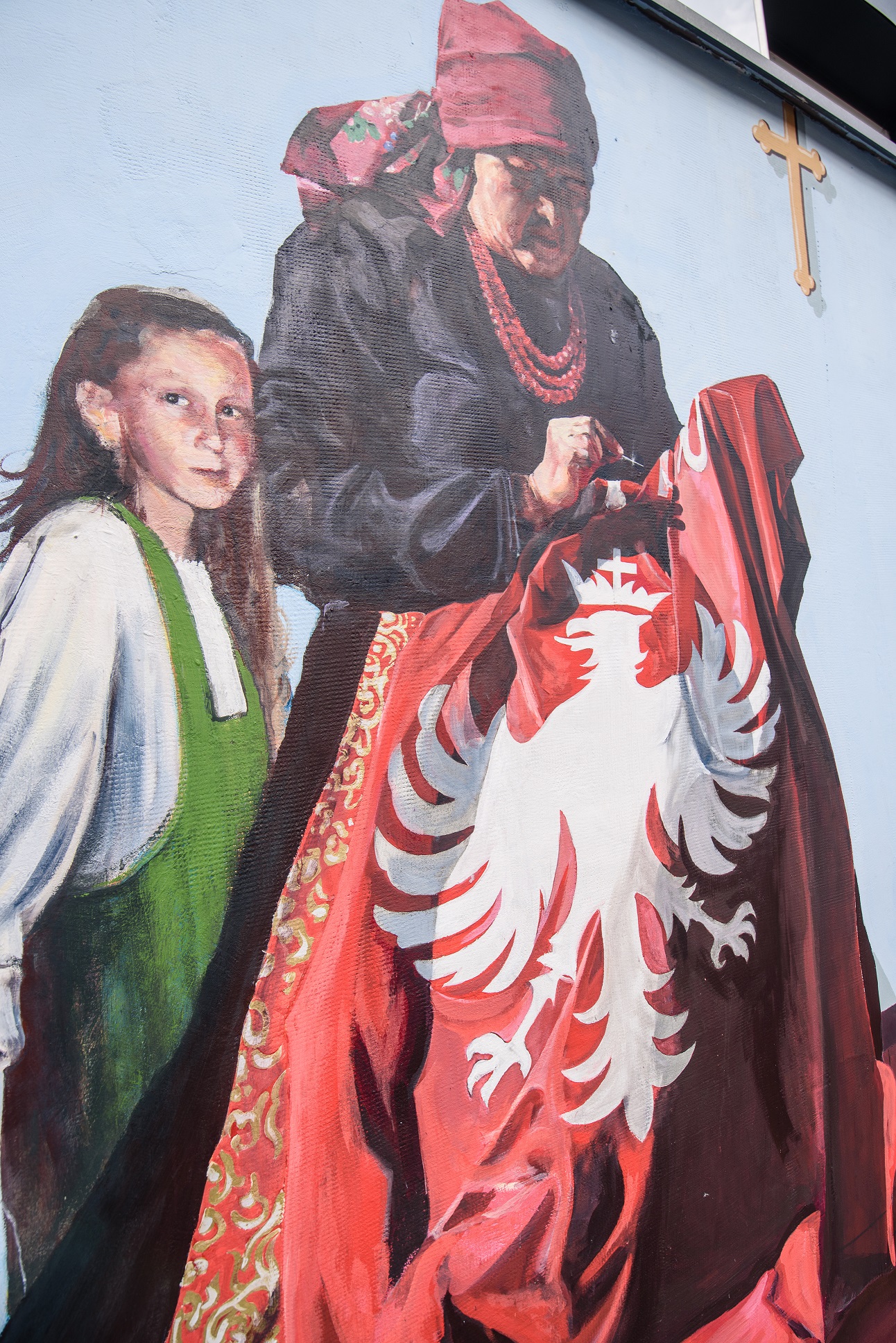 Kobieta w tradycyjnym śląckim stroju wyszywa orła na czerwonej tkaninie , towarzyszy jej dziewczynka twarzą zwrócona do oglądającego mural.