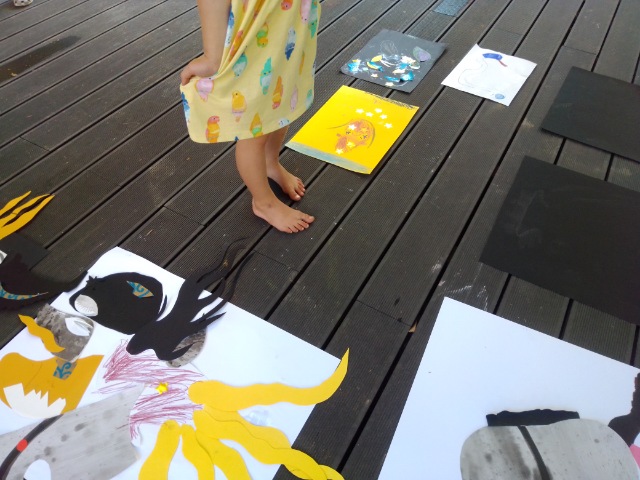 Zdjęcie przedstawia w lewym górnym rogu stopy dziewczynki i fragment jej żółtej letniej sukienki. Wokół dziewczynki wokół jej stup na podłodze leżą rozłożone prace namalowane przez dziewczynkę.
