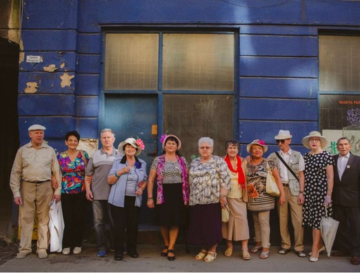 Na zdjęciu widać grupę jedenastu seniorów ubranych odświętnie, stojących na tle niebieskiej kamienicy.