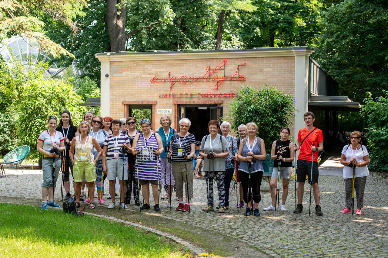 Zdjęcie przedstawia grupę uczestników warsztatów Nordic Walking stojących przed pawilonem Niepodległa. Miejsce spotkań.