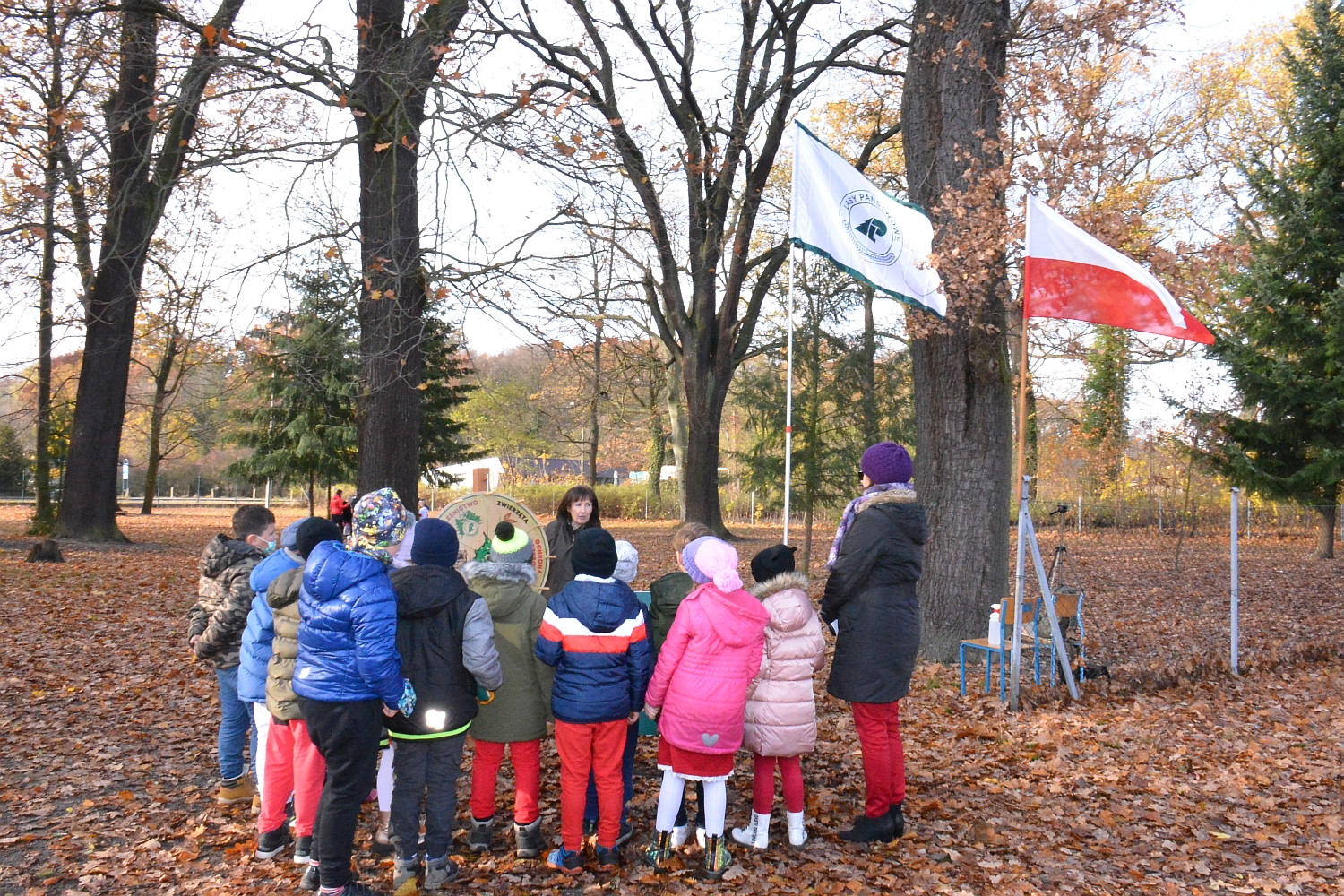 Na zdjęciu widoczna jest grupa dzieci z wychowawczynią podczas gry terenowej na terenie szkoły. Nad ich głowami powiewają flagi - flaga Polski oraz flaga Lasów Państwowych.