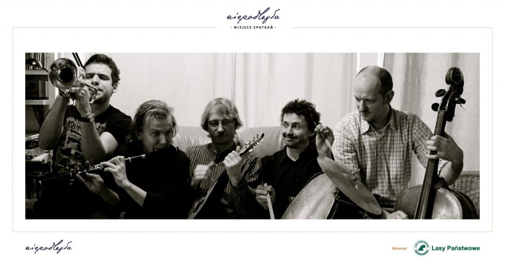 Fotografia przedstawia grupę 5 mężczyzn, którzy tworzą zespół Janusz Prusinowski Kompania. Każdy z nich trzyma instrument. Siedzą blisko siebie na kanapie. W tle regał z książkami, fotel wiklinowy i firanka.