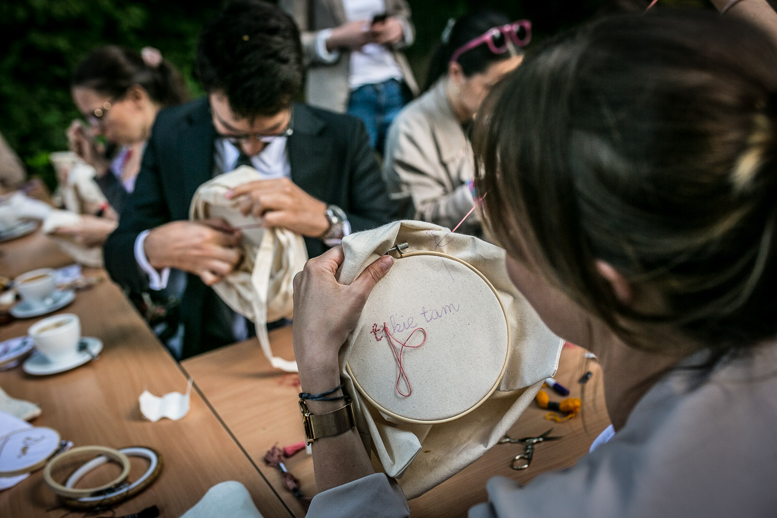 Zdjęcie przedstawia grupę osób siedzących przy stole w ogródku kawiarnianym, które haftują. Na blacie zostały ułożone przybory niezbędne do wykonania tych ręcznych prac.