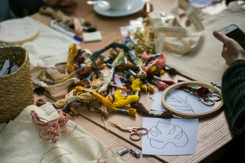 Zdjęcie przedstawia stół warsztatowy, na którym położone zostały materiały niezbędne do haftowania: muliny, nożyczki, szablony, bawełniane torby i tamborki.