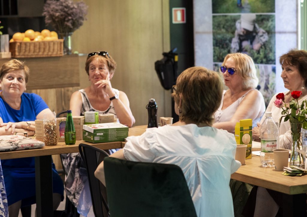 Zdjęcie zostało wykonane w pawilonie kulturalnym "Niepodległa. Miejsce Spotkań". Przedstawia grupę seniorów, siedzących przy stole i słuchających wykładu warsztatowego.